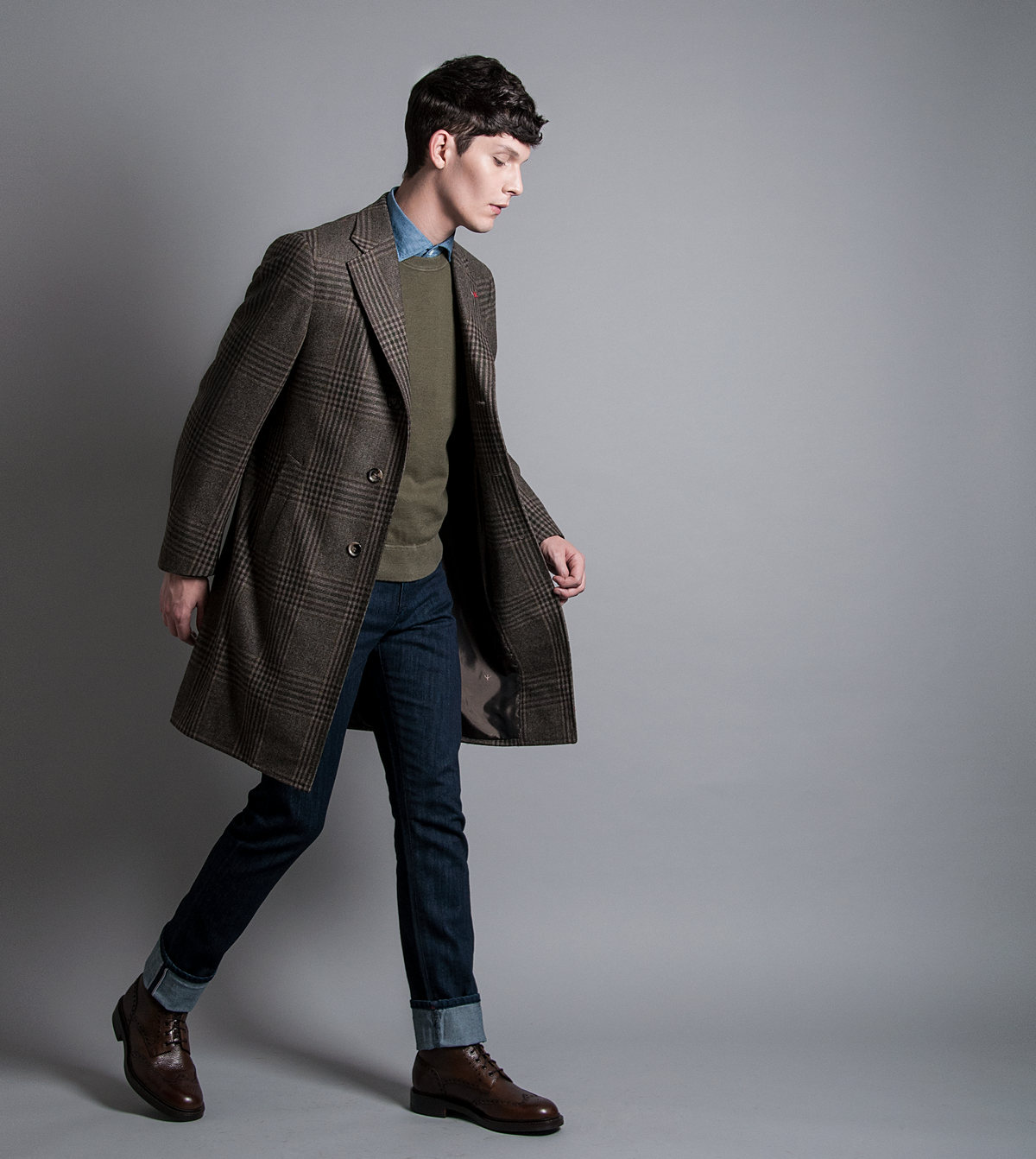 Modelo camina con abrigo a cuadros, suéter verde y jeans sobre fondo gris