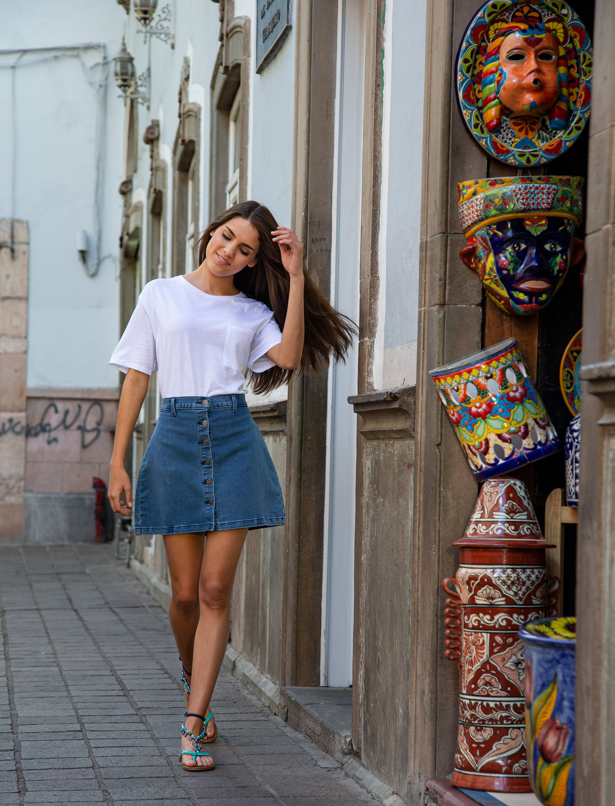 Modelo con playera blanca y falda de mezclilla camina junto a una tienda de artesanías mexicanas