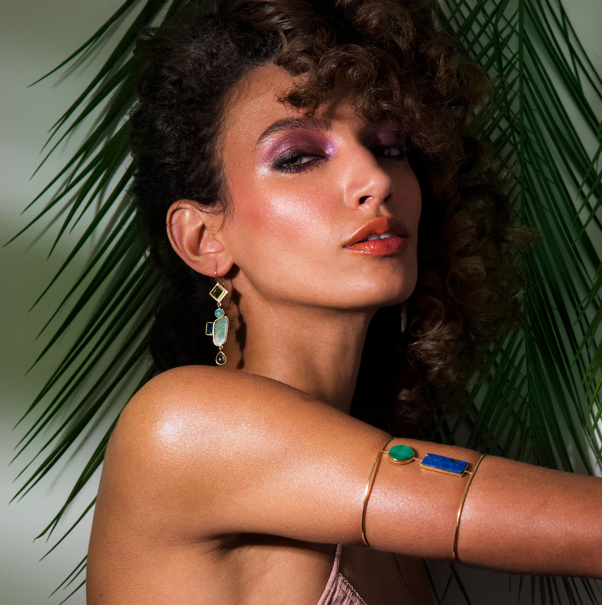 Curly-haired model with violet eyeshadow and a jeweled cuff among palm leaves