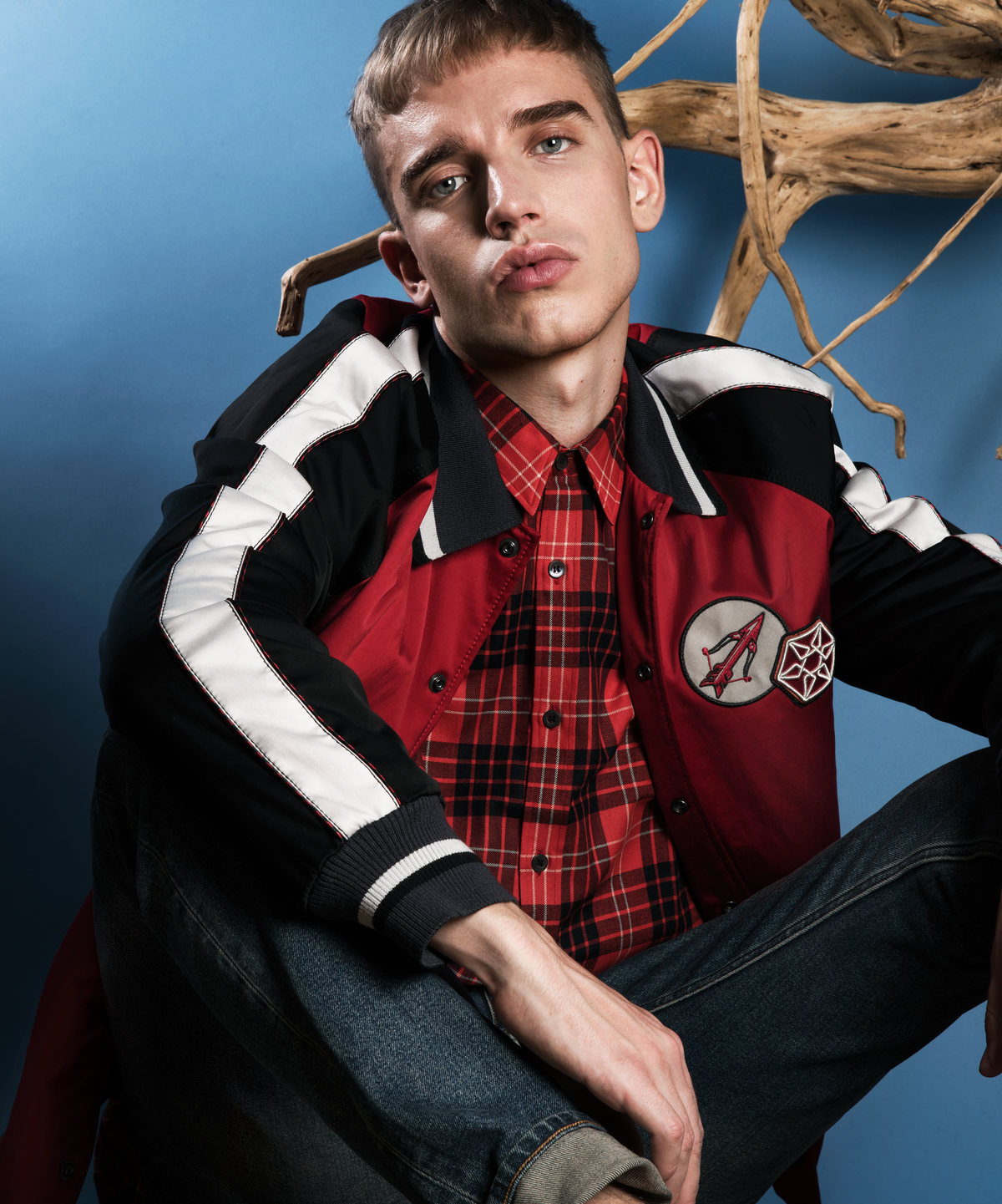 Model in a red varsity jacket and plaid shirt against blue with branches