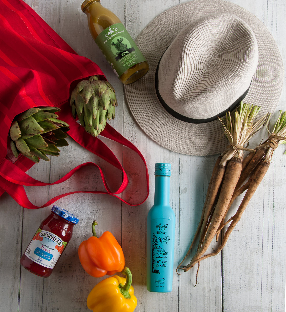 Red tote, straw hat, artichokes, carrots and peppers on white wood