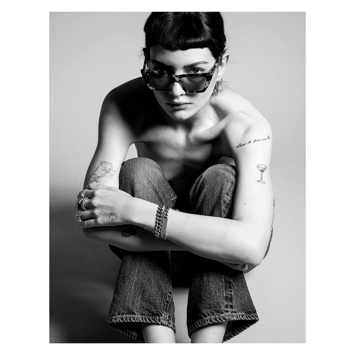 Black-and-white image of a model in sunglasses and jeans, seated hugging her knees