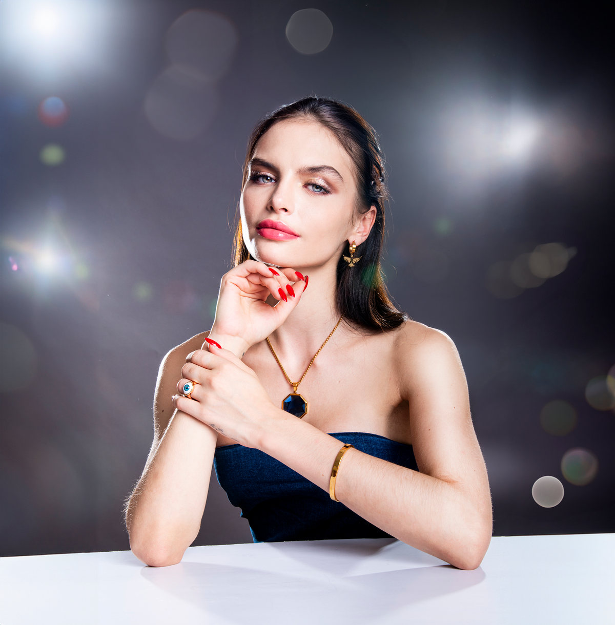 Model wearing a gold necklace and rings leaning on a table, dark background with lights