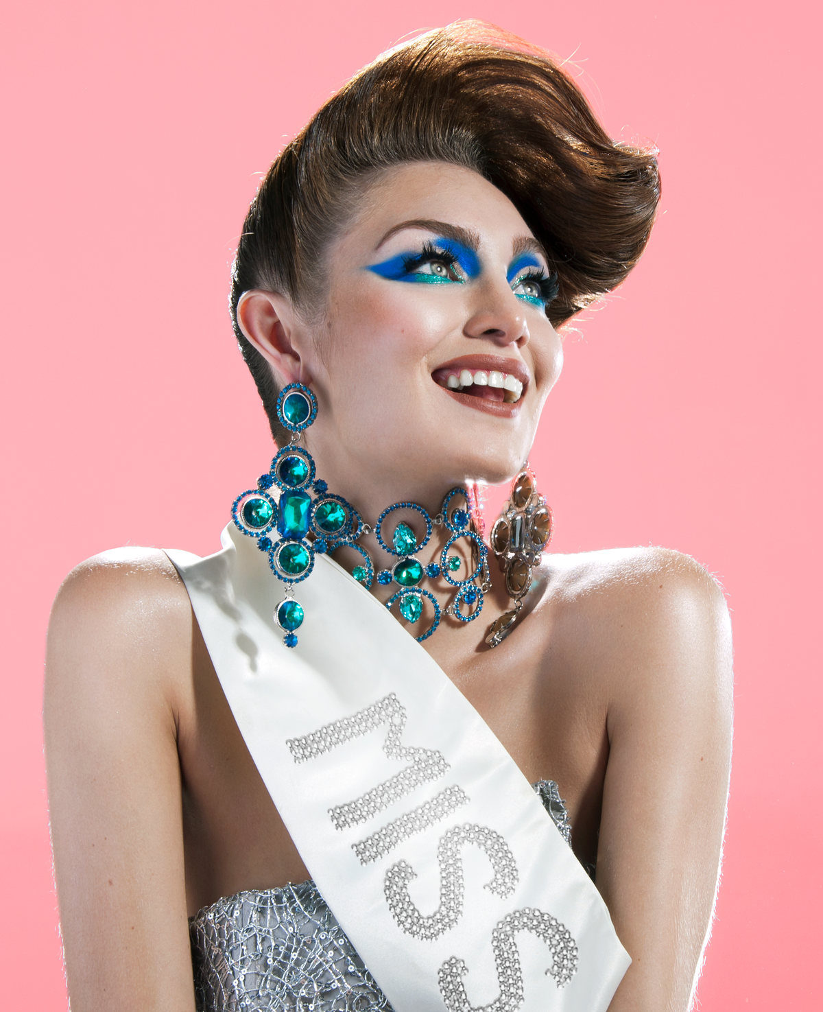Model wearing a pageant sash, blue eyeshadow and a turquoise necklace on pink