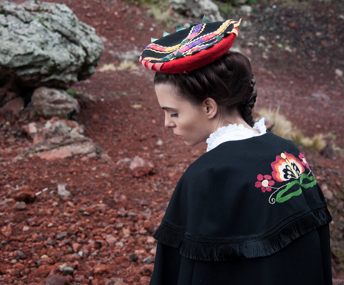 Modelo de espaldas con capa negra bordada y tocado de colores en un paisaje de roca volcánica
