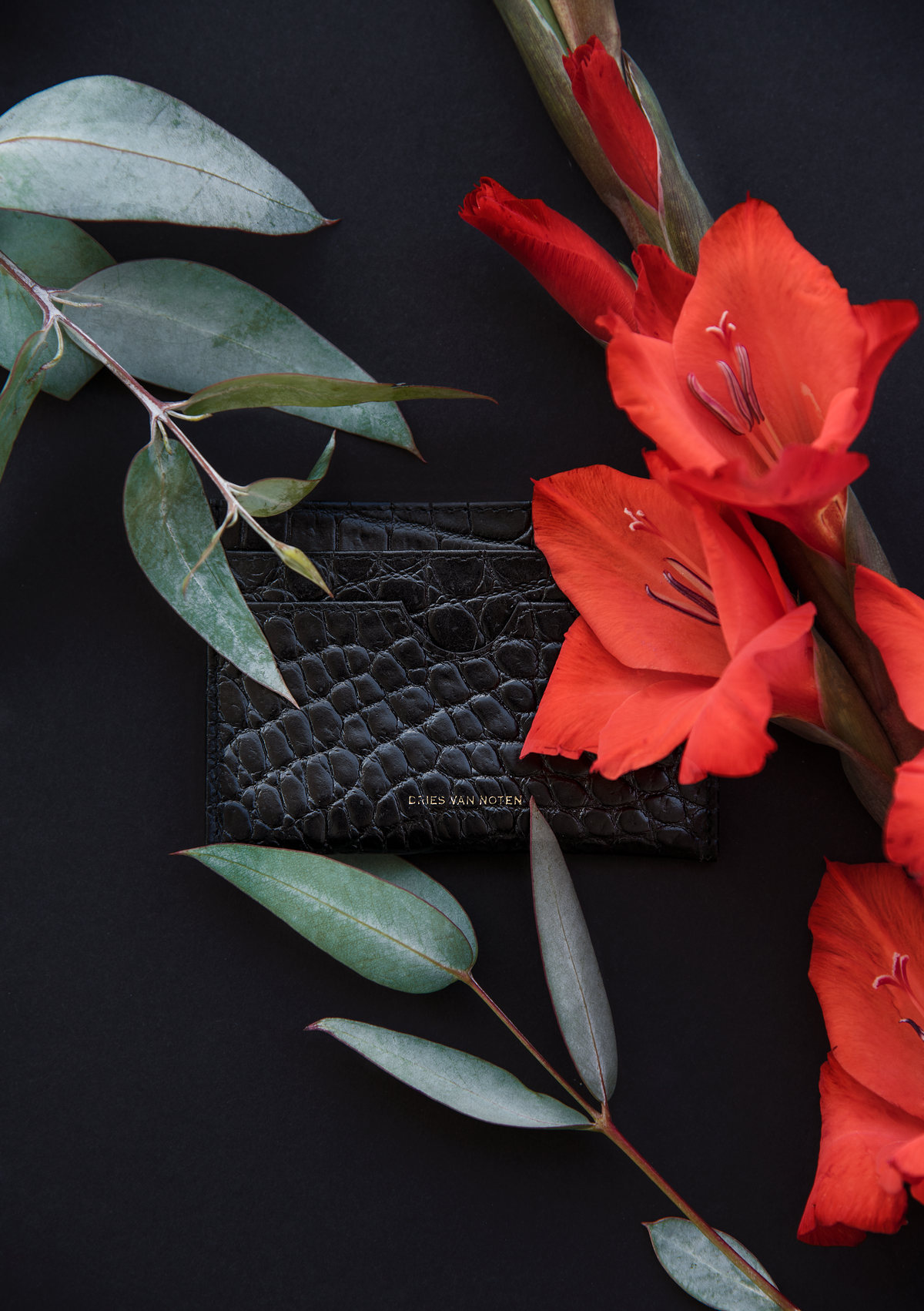 Cartera de piel negra texturizada entre gladiolas rojas y hojas sobre fondo negro