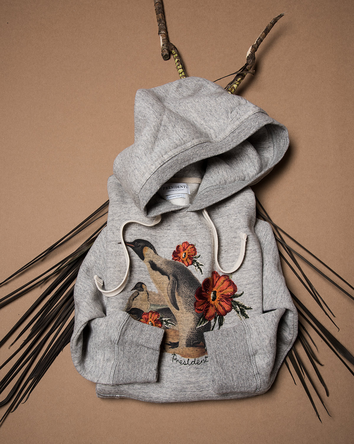 Sudadera gris con bordado floral colgada entre ramas secas, fondo café