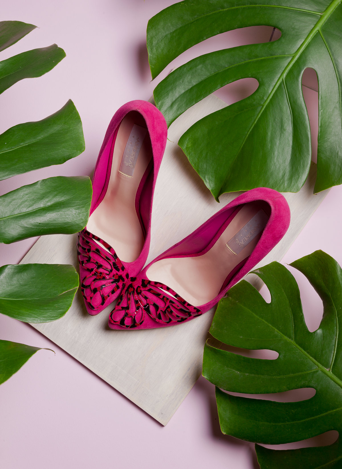 Zapatillas de gamuza fucsia con moño entre hojas de monstera sobre fondo rosa